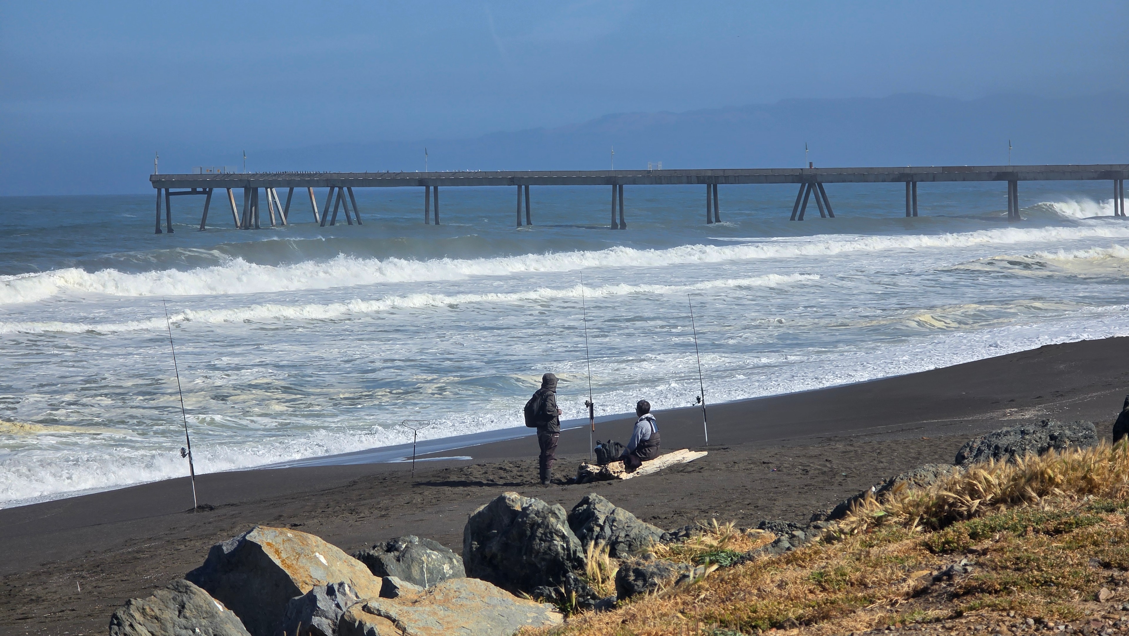 Fisherman sitting on a driftwood log along Pacifica's beach beneath the pier