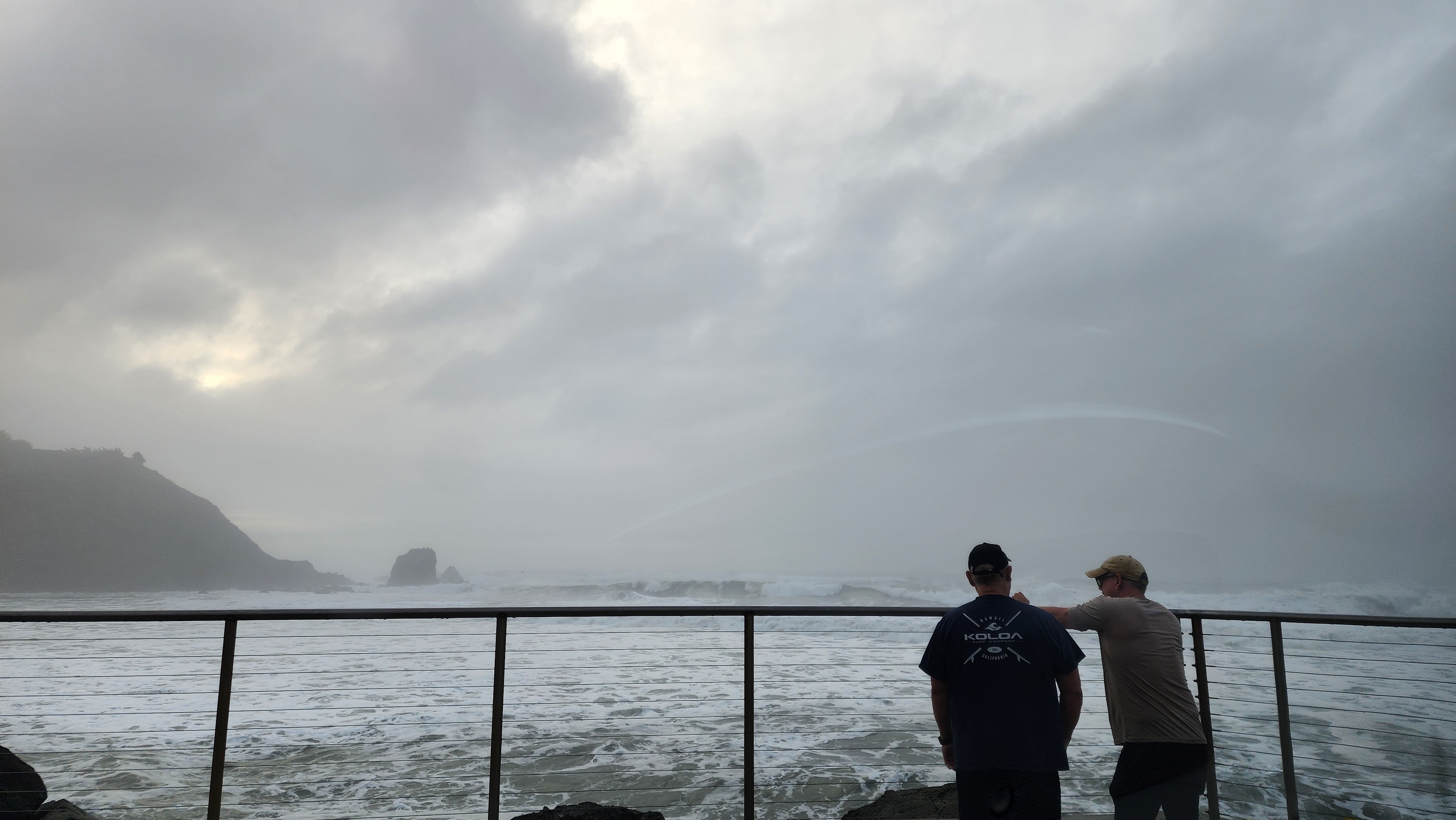People watching Pacifica's foggy coast from a railing beneath a rainbow